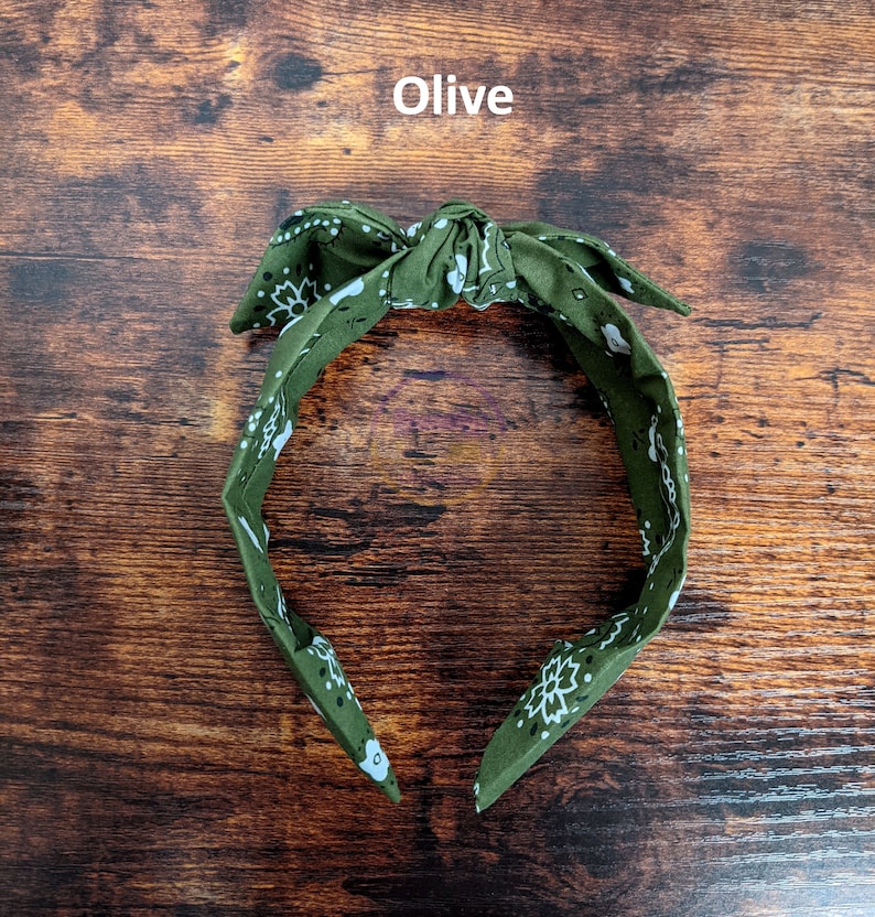 May include: Olive green bandana headband with a white paisley pattern. The headband is tied in a bow at the top.