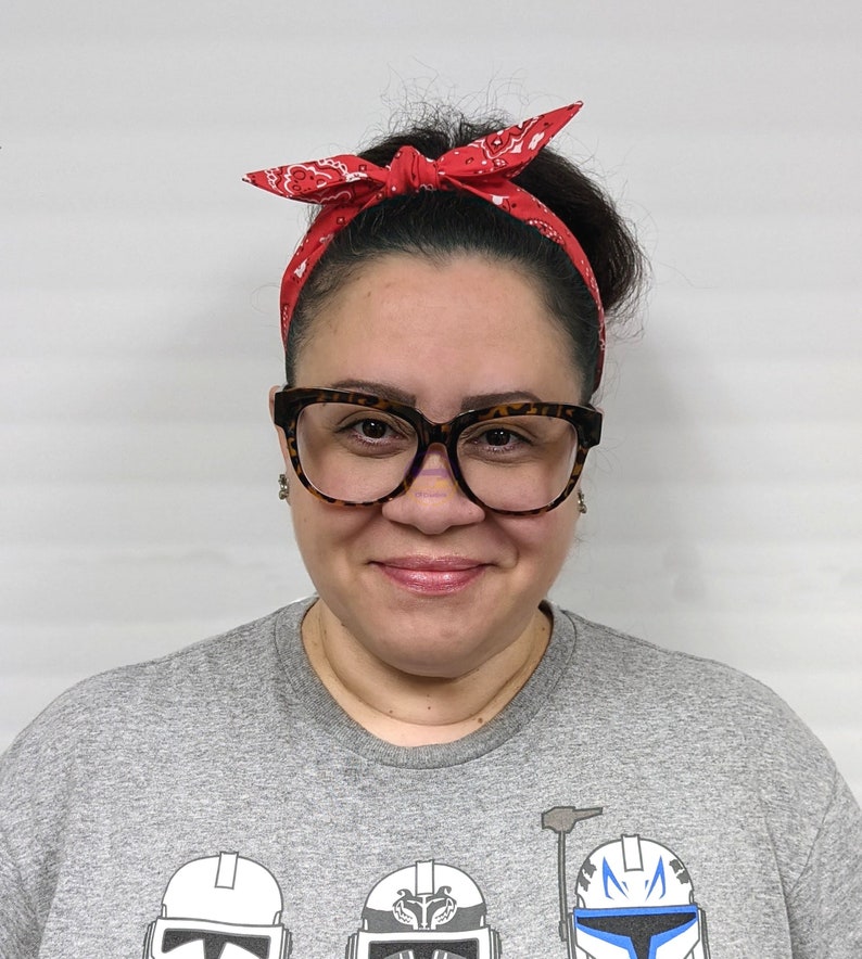 May include: A woman wearing a grey t-shirt with a Star Wars themed graphic print. She is wearing a red bandana headband and brown framed glasses.