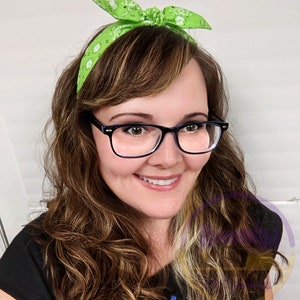 May include: A woman with long brown hair wearing a green bandana headband with white polka dots and black framed glasses. She is smiling and looking at the camera.