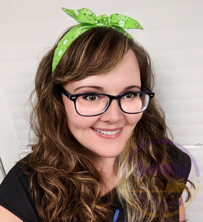 May include: A woman with long brown hair wearing a green bandana headband with white polka dots and black framed glasses. She is smiling and looking at the camera.