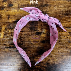May include: Pink bandana headband with a white paisley pattern. The headband is tied in a knot at the top.