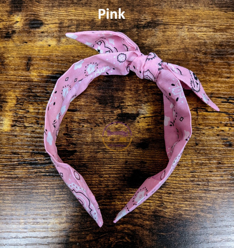 May include: Pink bandana headband with a white paisley pattern. The headband is tied in a knot at the top.