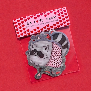 May include: A clear plastic package containing raccoon stickers. The raccoon is gray with black markings and a striped tail. The stickers are packaged with a pink header featuring red hearts and the text "DA LOVE PACK".