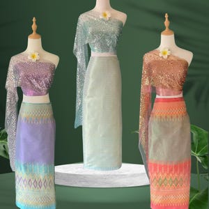 May include: Three traditional Thai outfits on mannequins. The outfits feature sequined tops and patterned skirts in shades of purple, seafoam green, and peach. Each mannequin is adorned with a flower. The outfits are displayed on a white platform against a green backdrop.