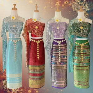 May include: Four traditional Thai outfits on mannequins. Each outfit features a long skirt, a top with a shawl, and a decorative belt. The outfits are in shades of blue, red, purple, and green, with gold accents and floral details.