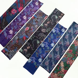 Könnte beinhalten: Eine Auswahl an farbenfrohen Batikstoffstreifen, die jeweils einzigartige Blumen- und geometrische Muster zeigen. Die Stoffe weisen eine Vielzahl von Farben auf, darunter Blau, Lila, Braun und Grün, mit aufwendigen Designs. Die Streifen sind diagonal angeordnet.