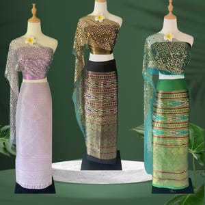 May include: Three traditional Thai outfits displayed on mannequins. The outfits feature ornate tops and long skirts in shades of purple, gold, and green. Each outfit is adorned with a flower and has a unique pattern and texture.