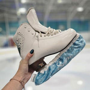 Könnte beinhalten: Weiße Eislaufschuhe mit einem blauen Samt-Schuhschutz. Die Schlittschuhe haben eine braune Ferse und weiße Schnürsenkel. Der Schuhschutz ist hellblau mit silbernen Akzenten. Die Schlittschuhe befinden sich auf einer weißen und blauen Oberfläche.