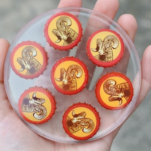 May include: A clear round coaster with six bottle caps embedded in the surface. Each bottle cap features a yellow and brown image of a ram's head with red trim.