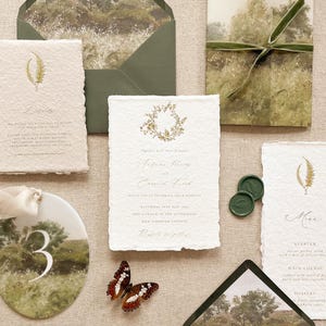 May include: Wedding stationery set with a nature theme. Includes invitations, menu cards, and an envelope with a landscape design. A butterfly and green wax seals are also present. The invitations have gold text and a botanical design.