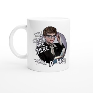 Puede incluir: Taza de cerámica blanca con una ilustración colorida de una mujer con gafas sosteniendo un libro. El texto en la taza dice "The clues are there. You a ho!"
