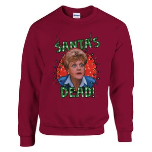 Puede incluir: Sudadera burdeos de cuello redondo con un gráfico que presenta la cara de una mujer rodeada de luces navideñas. El texto "SANTA'S DEAD!" está impreso encima y debajo de la imagen en letras verdes. La sudadera tiene mangas largas y cuello acanalado.