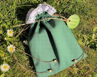 Puede incluir: Una bolsa verde oscuro con cordón y ribete de encaje blanco, sobre hierba con margaritas. La bolsa tiene una etiqueta que dice "Faerie of the Forest". El cordón es de cuerda con cuentas de madera.