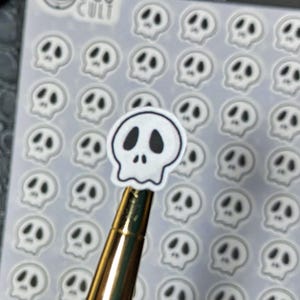 May include: A sheet of small, white skull stickers with black eye sockets and noses. One sticker is held up by a gold pen. The stickers are on a clear backing. The word "CULT" is visible in the top left corner.