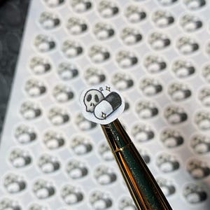 May include: Sheet of stickers featuring a skull and pill design. The stickers are small, round, and white with a black and gray illustration. A gold pen is holding one sticker, with a background of more stickers.