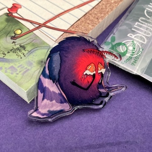 Peut inclure: Un charme en acrylique violet et rouge représentant une créature de dessin animé aux grands yeux et une expression triste. La créature a un corps rouge avec une aile rayée violette et blanche.