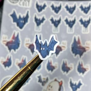 May include: A sheet of stickers featuring dark blue bat illustrations. One sticker is held by a gold pen. The bats have a watercolor effect, with a gradient of colours. The stickers are on a light purple background.