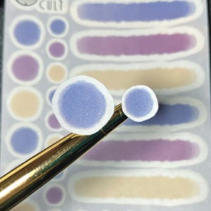 May include: Close-up of a sheet of stickers with a watercolor design. The stickers feature various shapes in shades of purple, blue, and beige. Two round stickers with a blue center are held by a gold-colored tool.