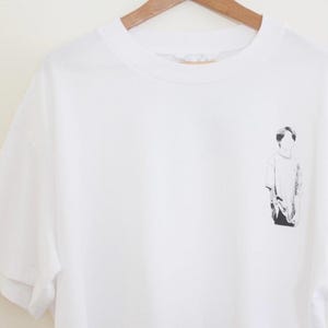 Puede incluir: Camiseta blanca con una ilustración en blanco y negro de una persona. La camiseta de manga corta está colgada de una percha de madera. La ilustración está en la parte delantera de la camiseta.