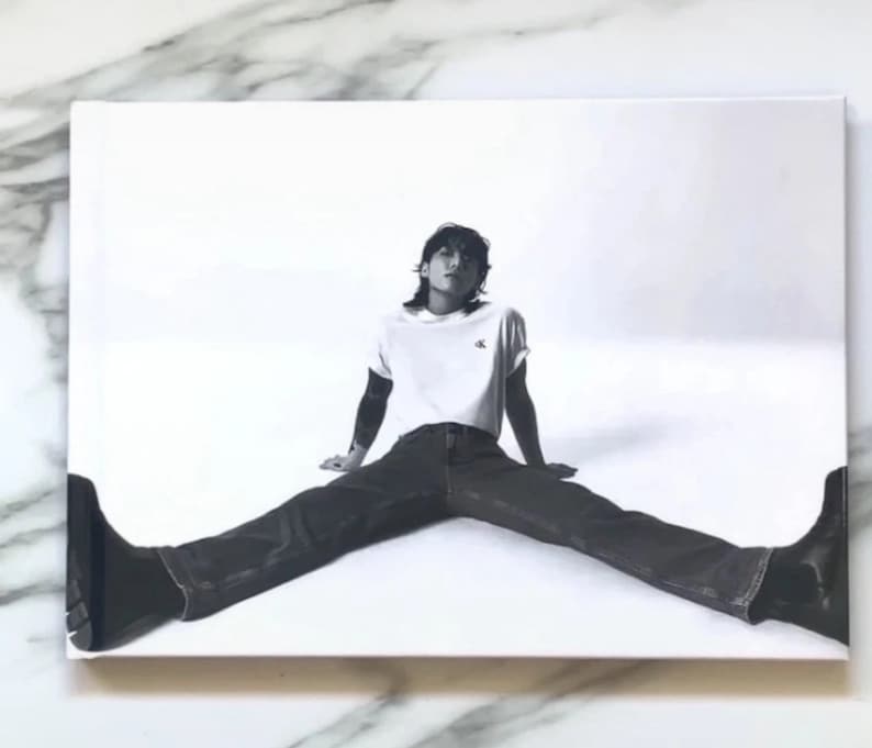 May include: A black and white photo of a person wearing a white t-shirt with a black logo, blue jeans, and black shoes. The person is sitting with their legs spread wide.