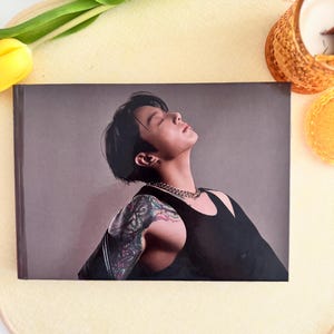 Puede incluir: Una impresión rectangular que presenta a una persona con cabello negro corto, un collar de cadena plateada y una camiseta sin mangas negra. La persona tiene un piercing en el labio y un tatuaje colorido en el brazo. El fondo es gris sólido.