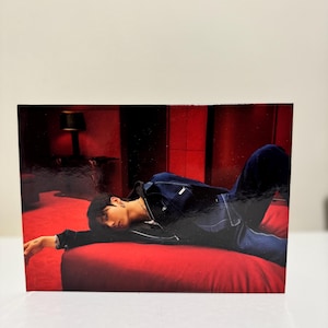 Può includere: Fotografia lucida con una persona in giacca e jeans blu scuro, sdraiata su un divano rosso. Lo sfondo è una stanza poco illuminata con pareti rosse e una lampada. La persona indossa una collana d'argento.