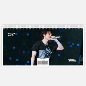 May include: A horizontal spiral-bound calendar featuring a photograph of a performer holding a microphone. The text 2027 appears on the left, and the name SUGA is printed on the right. A small colorland logo and barcode are centred at the bottom.