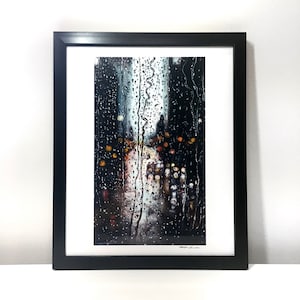 Peut inclure: Impression d'art encadrée représentant une scène de nuit pluvieuse. L'image montre une vue floue des lumières de la ville à travers une fenêtre striée de pluie. L'œuvre est encadrée dans un cadre noir avec un passe-partout blanc.