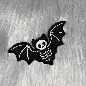Può includere: Toppa a forma di pipistrello ricamata in bianco e nero con un design a scheletro. Il pipistrello ha le ali spiegate e una testa a forma di teschio. La toppa è posta su uno sfondo di finta pelliccia grigia.