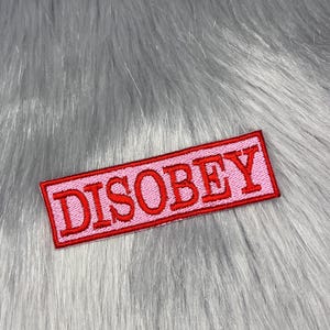 Puede incluir: Un parche rectangular rosa con la palabra "DISOBEY" bordada en rojo. El parche tiene un borde rojo y está colocado sobre un fondo de piel sintética gris.