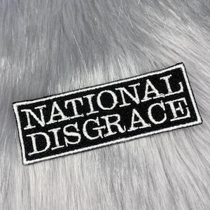 May include: A rectangular black and white patch with the words "NATIONAL DISGRACE" in white capital letters. The patch has a black border and is set against a backdrop of gray faux fur.