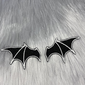 May include: Two black bat wing patches with white stitching details. The patches are displayed on a gray, fluffy surface. These patches are ideal for adding a spooky touch to clothing or accessories.