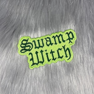 May include: A green embroidered patch with the words "Swamp Witch" in a gothic font. The patch has a lime green border and is set against a gray, furry background.
