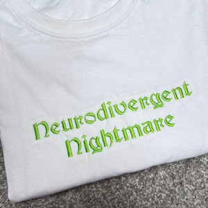 May include: White t-shirt with green embroidered text that reads "Neurodivergent Nightmare".