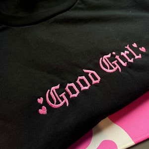May include: Black t-shirt with pink embroidered text that says "Good Girl" with two pink hearts on either side of the text.