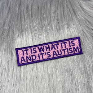 Può includere: Una toppa rettangolare ricamata con sfondo rosa chiaro, bordo viola scuro e testo viola scuro che recita IT IS WHAT IT IS AND IT'S AUTISM. La toppa presenta un bordo cucito pulito ed è centrata all'interno dell'inquadratura.