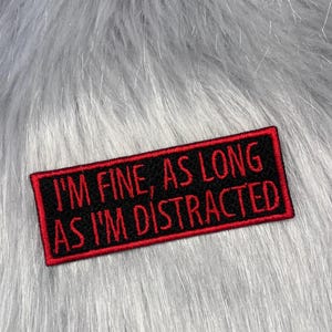 May include: A rectangular embroidered patch with a black background and a red border. The text I'M FINE, AS LONG AS I'M DISTRACTED is stitched in red capital letters across the centre of the patch, featuring a clean, bold, sans-serif font style.
