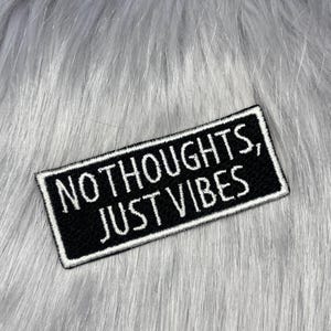 May include: A rectangular black embroidered patch with a white border and white stitched text that reads, "NO THOUGHTS, JUST VIBES." The patch features a clean, bold font and is designed for application onto fabric surfaces as a decorative accessory.