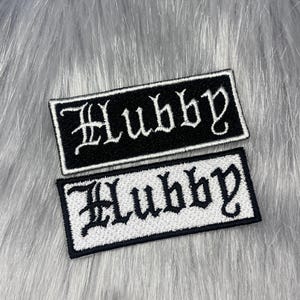 Könnte beinhalten: Zwei rechteckige gestickte Aufnäher mit dem Wort Hubby in einer Schriftart im gotischen Stil. Ein Aufnäher hat weiße Buchstaben auf schwarzem Hintergrund, der andere schwarze Buchstaben auf weißem Hintergrund mit schwarzem Rand.