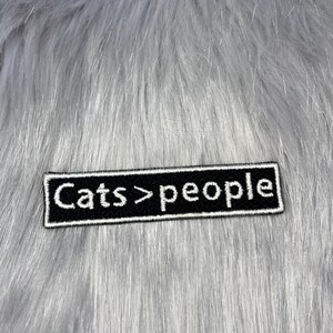 May include: A rectangular black embroidered patch with a white border and white stitched text that reads "Cats > people". The patch features a clean, bold font and is designed for application onto fabric surfaces as a decorative accessory.