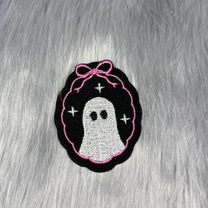 Puede incluir: Un parche bordado que presenta un fantasma blanco centrado sobre un fondo ovalado negro. El diseño está enmarcado por un borde cosido en rosa con un lazo rosa en la parte superior y pequeñas estrellas blancas de cuatro puntas.