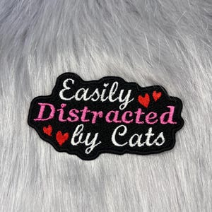 May include: A black embroidered patch with a scalloped edge featuring the white text Easily and Cats, with the pink text Distracted in the centre. Four small red hearts are interspersed throughout the text on the black fabric background.