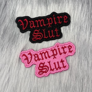 Könnte beinhalten: Zwei gestickte Aufnäher mit dem Text Vampire Slut in einer gotischen Schriftart. Ein Aufnäher hat einen schwarzen Stoff mit roter Stickerei, der andere einen rosa Stoff mit dunkelrosa Stickerei. Beide haben eine gestanzte Form um den Text.