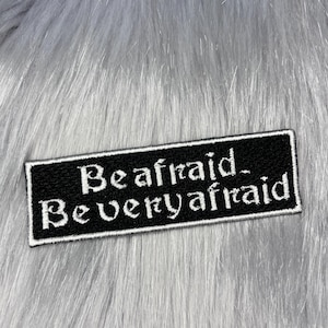 May include: A rectangular black embroidered patch with a white border and white text that reads Be afraid. Be very afraid. The patch features a gothic-style font and is positioned against a background of long, soft, light grey synthetic fur.