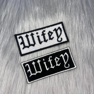 Könnte beinhalten: Zwei rechteckige bestickte Aufnäher mit dem Wort Wifey in einer Schriftart im gotischen Stil. Ein Aufnäher hat einen weißen Hintergrund mit schwarzer Schrift und schwarzem Rand, der andere einen schwarzen Hintergrund mit weißer Schrift und weißem Rand.