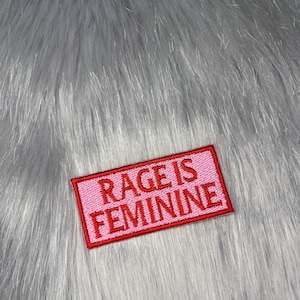 Puede incluir: Un parche rectangular rosa con las palabras "RAGE IS FEMININE" bordadas en rojo. El parche está sobre un fondo de piel sintética gris.