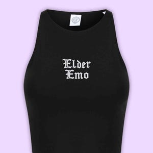 Può includere: Un top corto nero senza maniche con scollo alto e la scritta Elder Emo stampata in bianco con un carattere in stile gotico al centro del petto. Un'etichetta bianca del marchio con le lettere SF è visibile all'interno dello scollo.