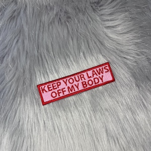 May include: A rectangular embroidered patch with a light pink background and a red border. The text KEEP YOUR LAWS OFF MY BODY is stitched in red capital letters across the centre of the patch, which features a clean, stitched edge finish.