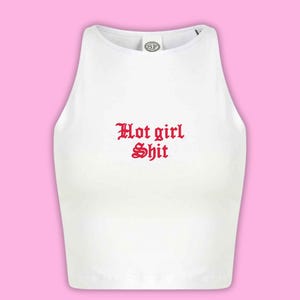 May include: White crop top with the text "Hot girl Shit" in red gothic font. The top has a scoop neck and a racerback design. The background is a solid pink colour.
