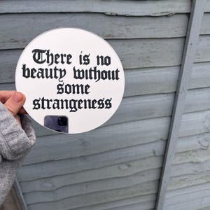 May include: A white round mirror with a black gothic font quote that reads "There is no beauty without some strangeness".